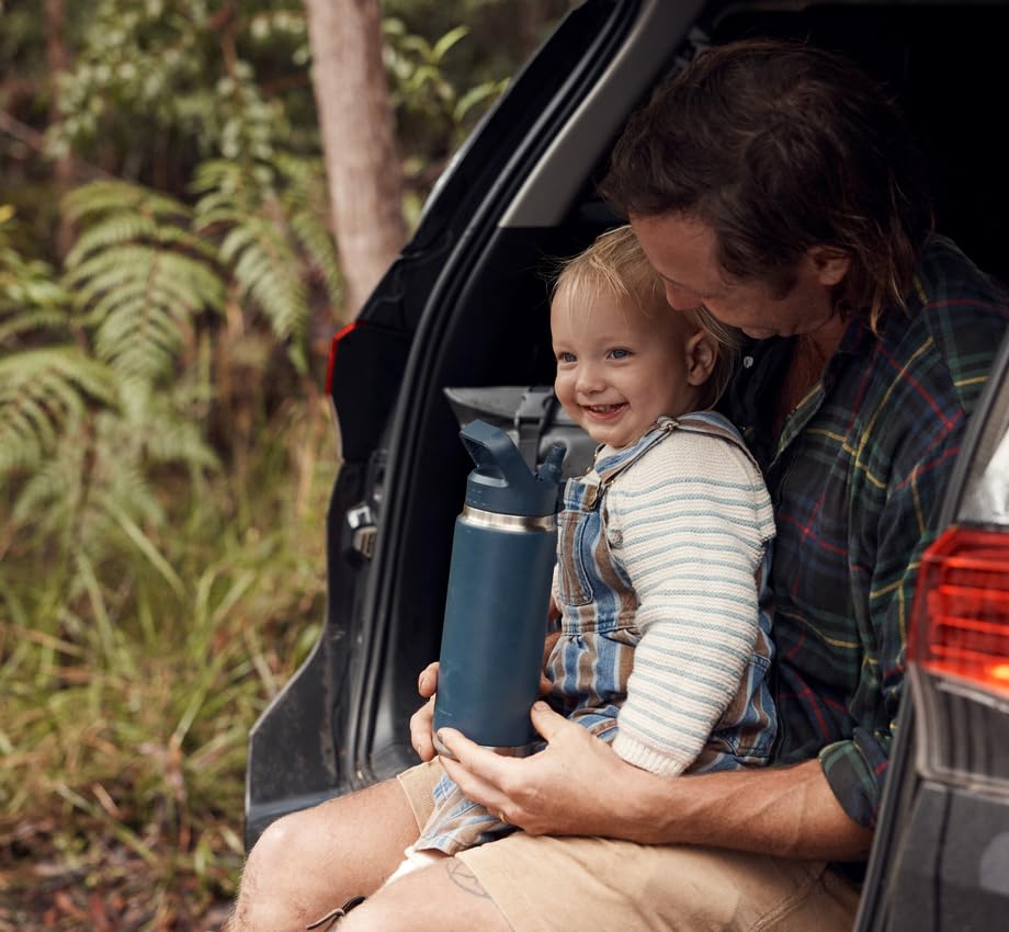 Man and child sitting in a car with a blue thermos, surrounded by greenery
