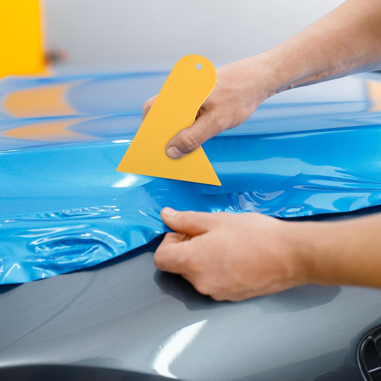 Person applying blue car wrap with a yellow tool on a car hood.
