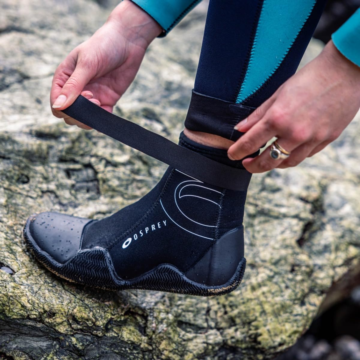 Person adjusting a black wetsuit boot on a rocky surface