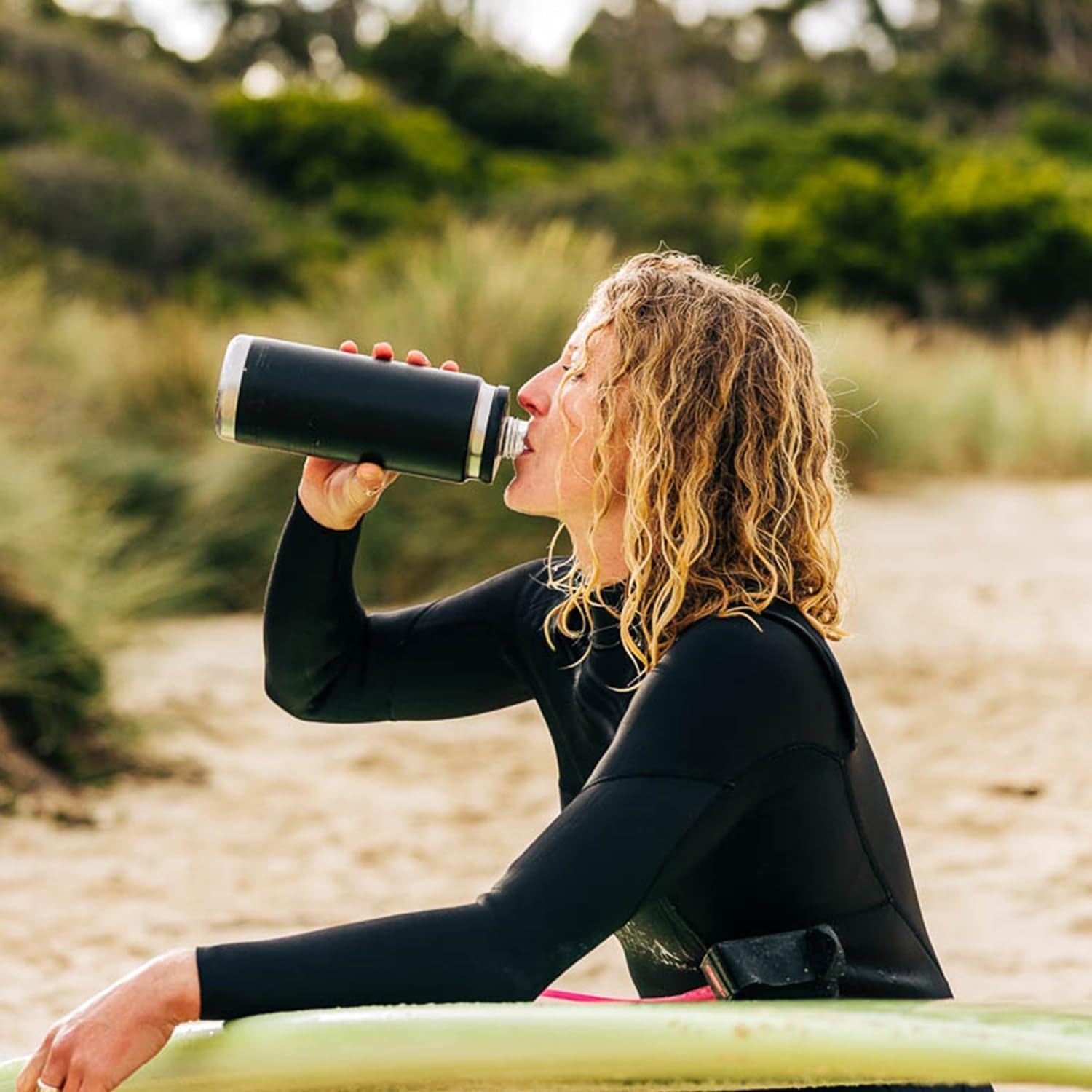 Person in a wetsuit drinking from a insulated bottle on a beach.