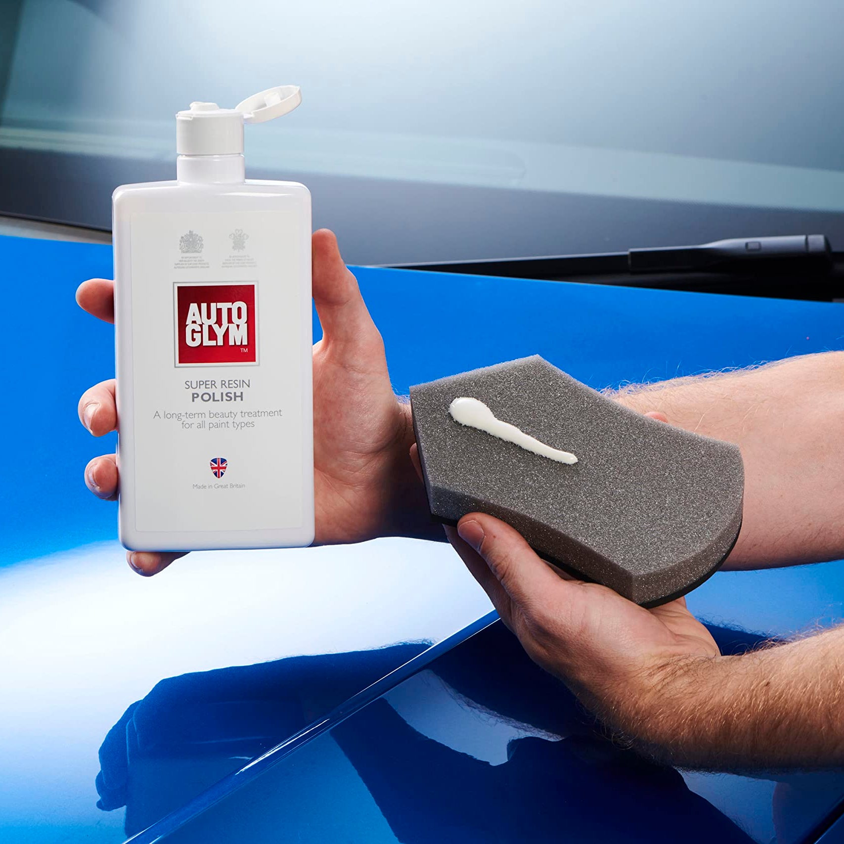 Person holding a bottle of AUTO GLAM car polish and a foam pad in front of a blue car.