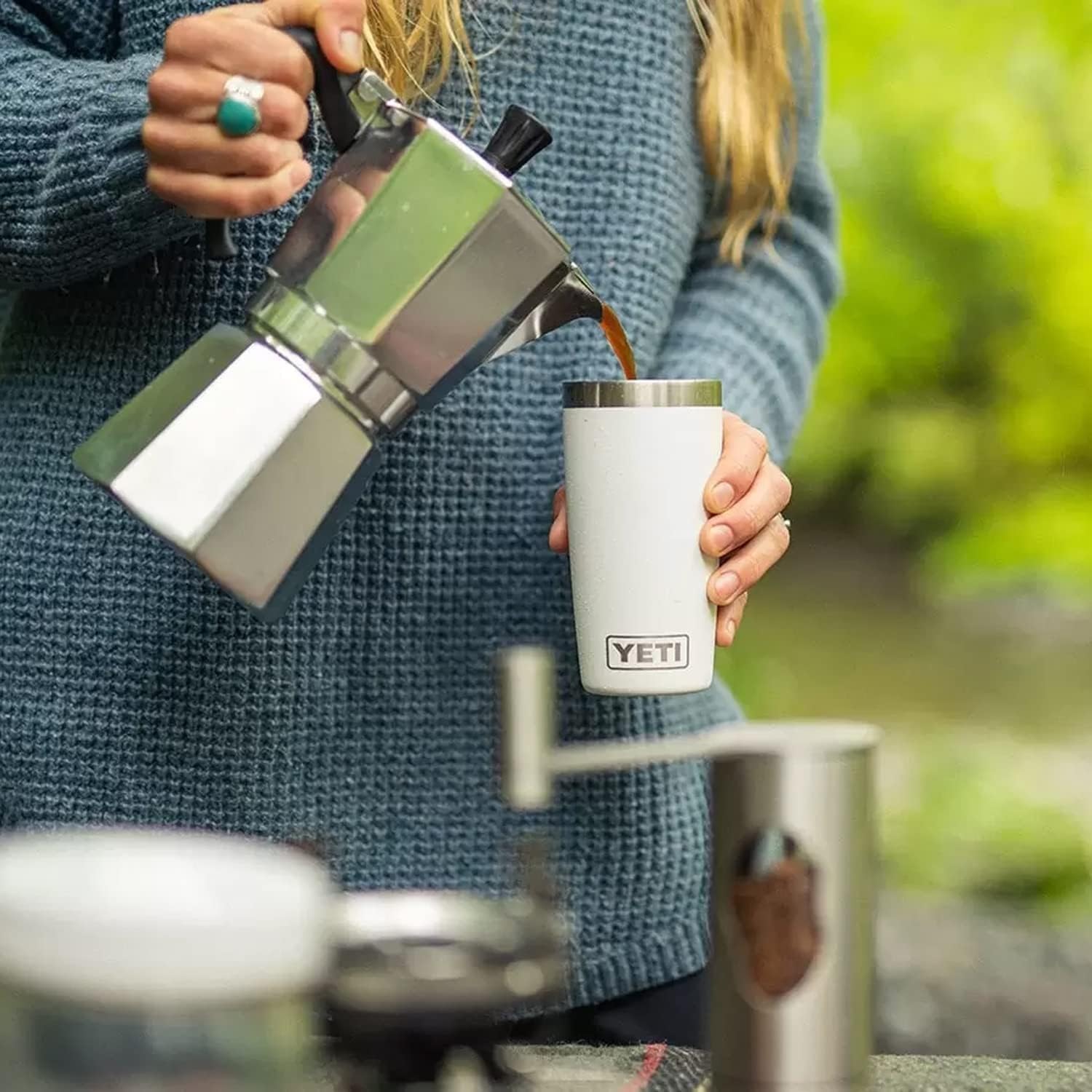 Person pouring coffee from a French press into a YETI mug outdoors.
